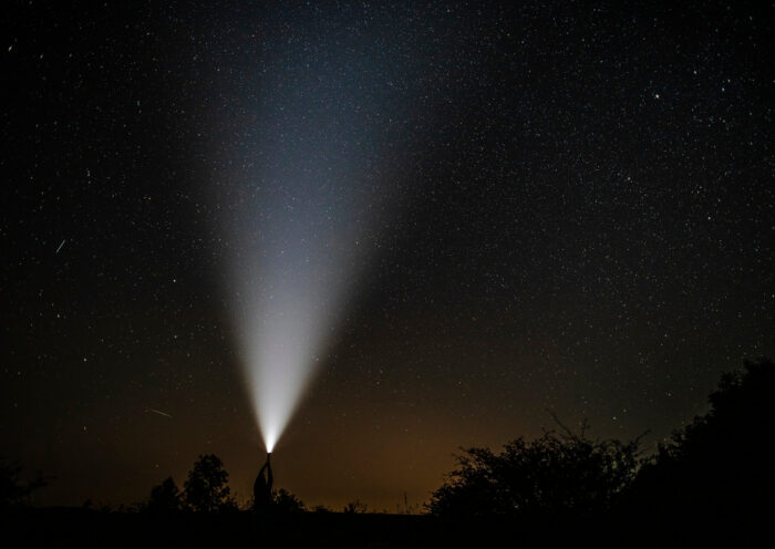 Nachtaufnahme eines hellen Suchscheinwerferstrahls, der in den sternenklaren Himmel leuchtet, mit Silhouetten von Bäumen und einem Kirchturm im Vordergrund – symbolisiert Orientierung und Vorbereitung in dunklen oder ungewissen Zeiten.