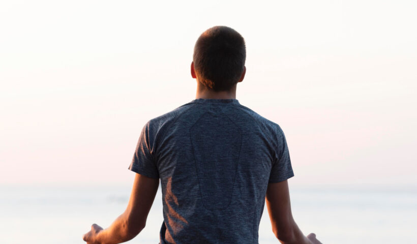 Person in Meditationshaltung im Lotussitz auf einem Holzsteg am Meer während des Sonnenuntergangs, von hinten fotografiert – symbolisiert Achtsamkeit, innere Ruhe und mentale Stärke als Teil der Krisenpsychologie und Stressbewältigung.