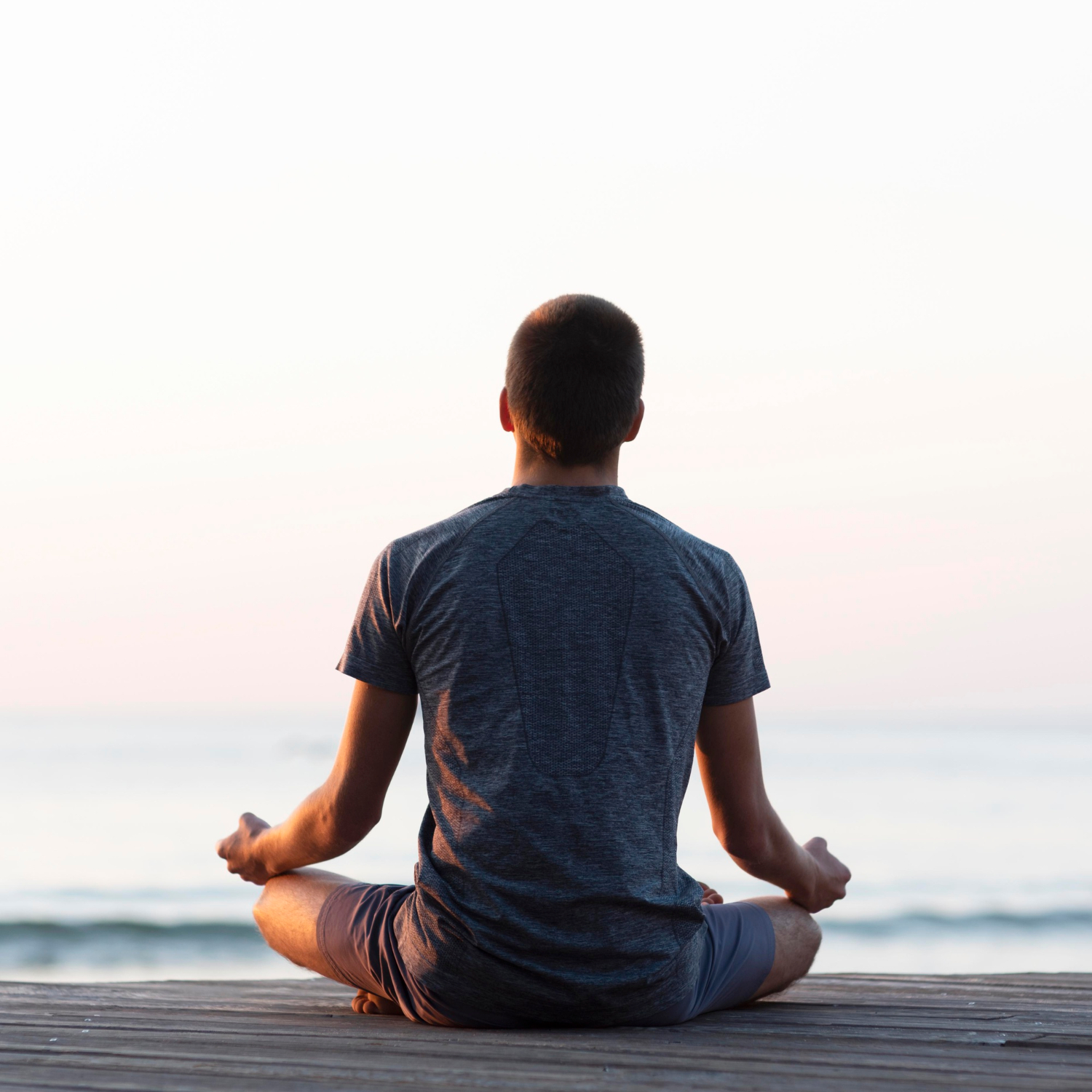 Person in Meditationshaltung im Lotussitz auf einem Holzsteg am Meer während des Sonnenuntergangs, von hinten fotografiert – symbolisiert Achtsamkeit, innere Ruhe und mentale Stärke als Teil der Krisenpsychologie und Stressbewältigung.