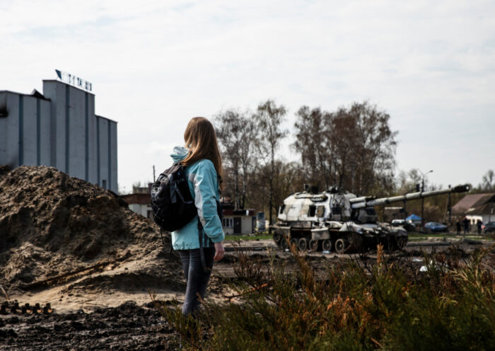 Jugendliche mit Rucksack steht auf einem verwüsteten Gelände mit aufgeworfener Erde und Schutt, während im Hintergrund ein zerstörter Panzer und ein Industriegebäude zu sehen sind – symbolisiert die Auswirkungen von Krieg und Zerstörung auf den Alltag und die Bedeutung von Frieden.