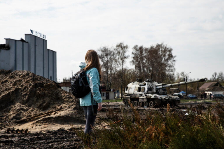 Jugendliche mit Rucksack steht auf einem verwüsteten Gelände mit aufgeworfener Erde und Schutt, während im Hintergrund ein zerstörter Panzer und ein Industriegebäude zu sehen sind – symbolisiert die Auswirkungen von Krieg und Zerstörung auf den Alltag und die Bedeutung von Frieden.