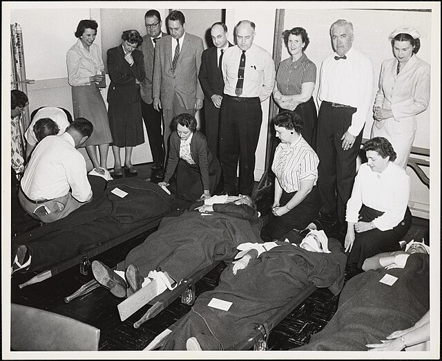 Historisches Schwarz-Weiß-Foto einer Gruppe von Personen während einer Zivilschutz- oder Erste-Hilfe-Übung in den 1950er oder 1960er Jahren. Mehrere Personen üben die Versorgung von Verletzten auf Tragebahre-Liegen, während andere zuschauen und anleiten. Die Szene zeigt die Bedeutung von strukturierter Ausbildung und Gemeinschaft im Katastrophenschutz.
