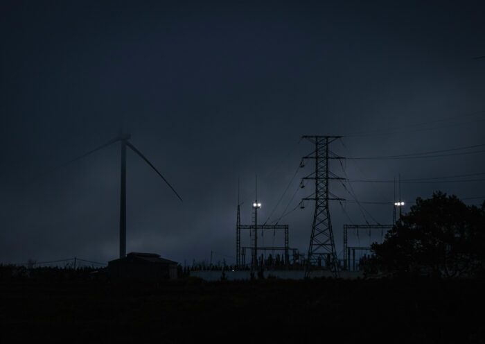 Dämmerungsaufnahme eines Strommasts und einer Windkraftanlage in der Abenddämmerung, beleuchtet von wenigen Scheinwerfern, mit dunklem Himmel und Silhouetten von Bäumen im Vordergrund – symbolisiert die Abhängigkeit von Energieinfrastruktur und die Bedeutung von Notfallvorsorge bei Stromausfällen.