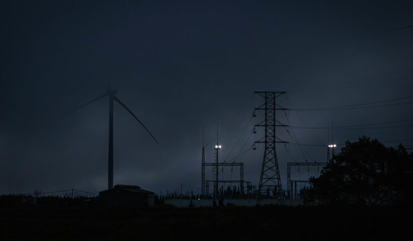 Dämmerungsaufnahme eines Strommasts und einer Windkraftanlage in der Abenddämmerung, beleuchtet von wenigen Scheinwerfern, mit dunklem Himmel und Silhouetten von Bäumen im Vordergrund – symbolisiert die Abhängigkeit von Energieinfrastruktur und die Bedeutung von Notfallvorsorge bei Stromausfällen.
