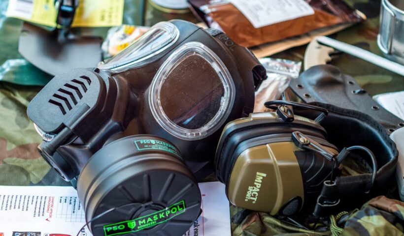 Notfallausrüstung mit zwei Schutzmasken (eine Gasmasken-Atemschutzmaske der Marke MaskPol und ein Gehörschutz der Marke Impact Sport) auf einem Tarnmuster-Stoff, umgeben von weiteren Überlebensausrüstungsgegenständen wie einem Notizbuch, Klebeband, Metallbehältern und einem Clipboard. Die Szene zeigt die Vorbereitung auf mögliche Krisensituationen.