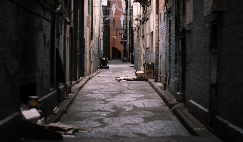 Dunkle, enge Gasse in einer städtischen Umgebung mit verwitterten Backsteinmauern, verstreutem Müll, nassen Pflastersteinen und geschlossenen Rollläden – Symbolbild für urbane Herausforderungen und Sicherheitsrisiken in Städten.